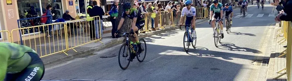 Cyclists racing on a street lined with spectators behind yellow barriers on a sunny day.
