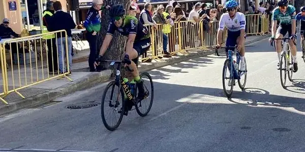 Cyclists racing on a street lined with spectators behind yellow barriers on a sunny day.