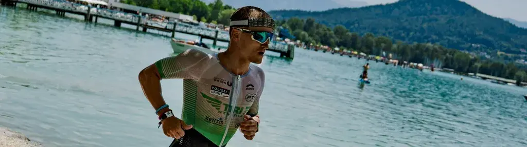 Athlete running by a lake, wearing a triathlon suit and sunglasses, with mountains and trees in the background.