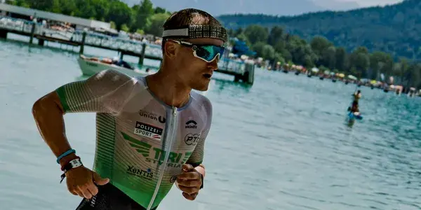 Athlete running by a lake, wearing a triathlon suit and sunglasses, with mountains and trees in the background.