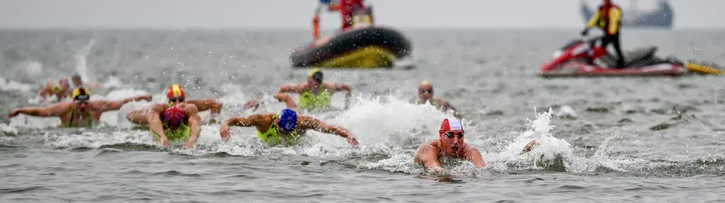 Swimmers in colorful caps compete in open water, with support boats in the background on a cloudy day.