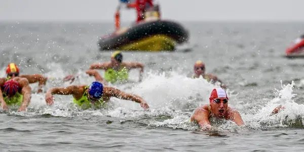 Swimmers in colorful caps compete in open water, with support boats in the background on a cloudy day.